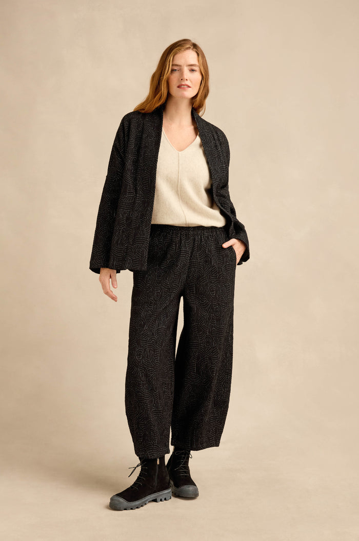 A woman stands against a beige background wearing the Stitched Jacquard Jacket in a boxy silhouette, paired with a cream V-neck top and black boots, with one hand in her pocket.