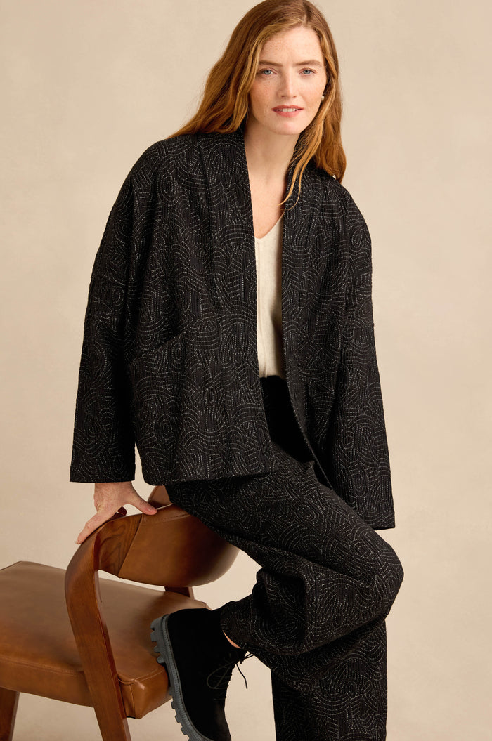 A person with long hair sits on a wooden chair against a plain beige background, wearing the Stitched Jacquard Jacket and matching pants, paired with a light-colored top and black boots. The jacket’s boxy silhouette lends modern structure to the ensemble.