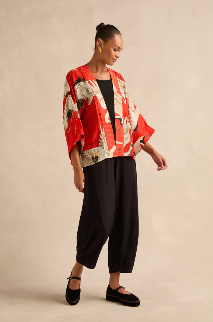 A woman stands against a neutral backdrop wearing the red Stork Kimono, featuring an Art Deco crane pattern, paired with a black top, loose black pants, and black shoes.