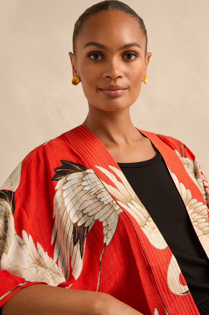 A woman with a neutral expression wears the Stork Kimono, featuring red floral patterns, over a black top and gold earrings against a light background.