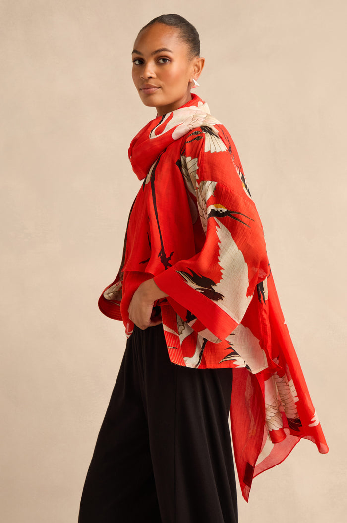 A woman stands against a beige background, wearing a red floral kimono-style top and black pants, her look elegantly finished with the Stork Scarf draped loosely around her neck as she looks at the camera.