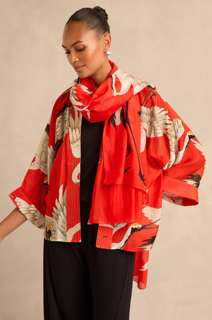 A woman stands against a beige background, wearing a red patterned kimono-style jacket and the Stork Scarf featuring white cranes, draped over a black outfit.