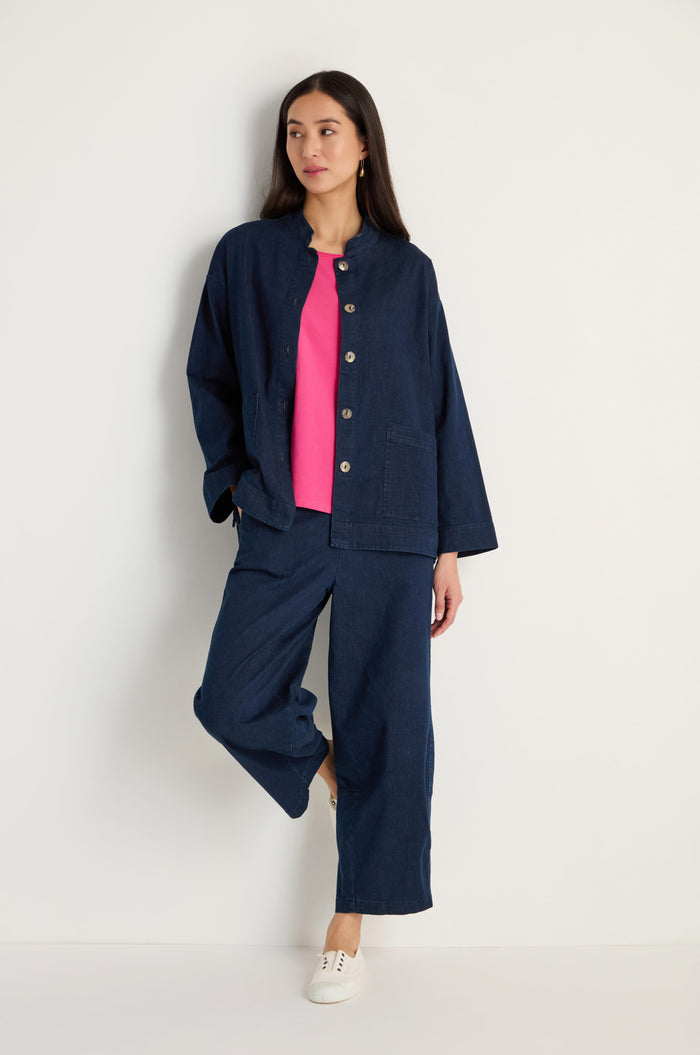 A woman leans against a white wall wearing the Stretch Denim Boxy Jacket and matching pants, paired with a pink top and white shoes.