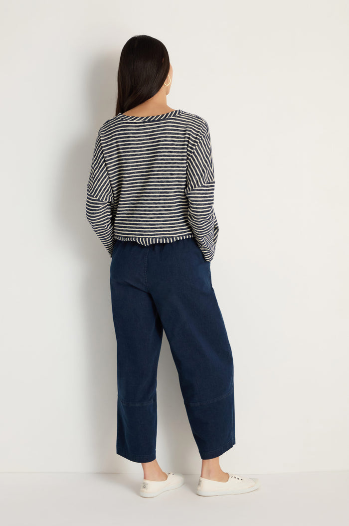 A person with long dark hair, wearing a black and white striped long-sleeve shirt, Stretch Denim Bubble Trousers in navy, and white sneakers stands facing a white wall with their back to the camera.
