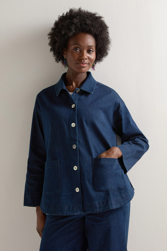 An individual with an afro hairstyle wears a Stretch Denim Jacket in dark blue with white buttons, colorful earrings, and a boxy fit, layered against a simple background.