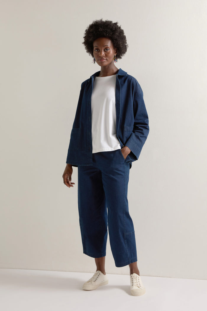 A person stands against a plain background, showcasing a boxy silhouette in the Stretch Denim Jacket layered over a white shirt, paired with matching pants and crisp white sneakers.