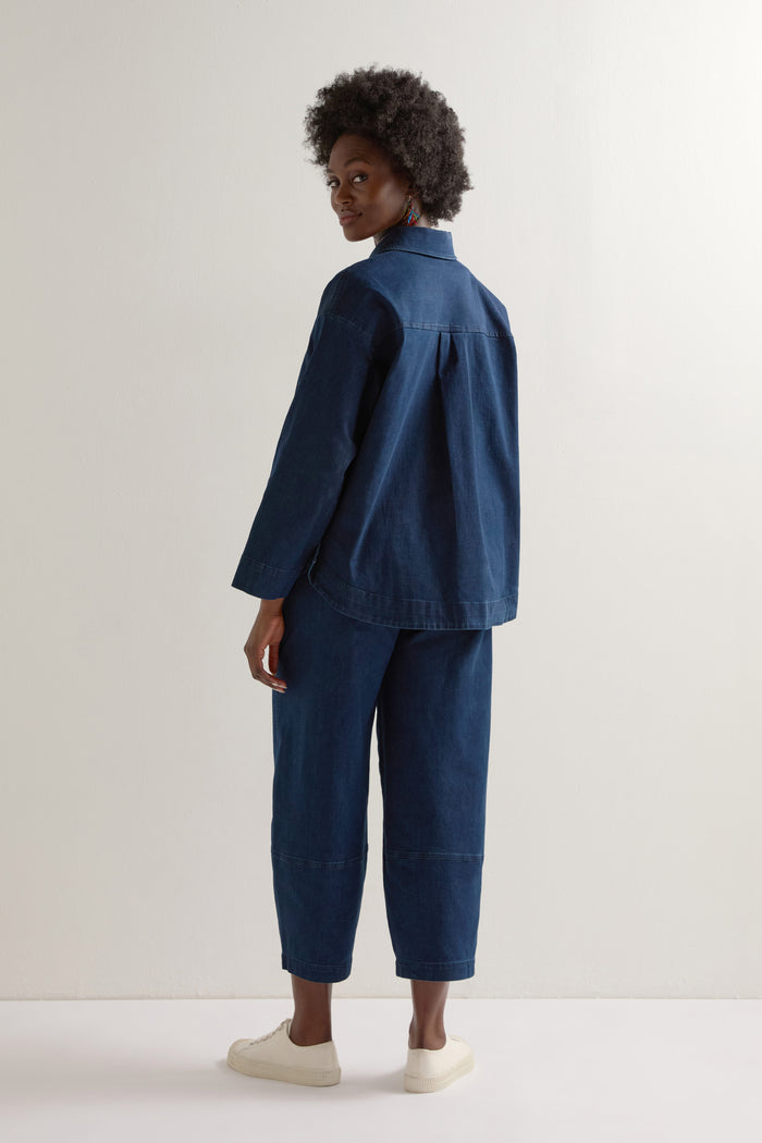 Dressed in a Stretch Denim Jacket, a person exudes effortless style with its boxy silhouette, complemented by denim pants and white sneakers against a simple backdrop.