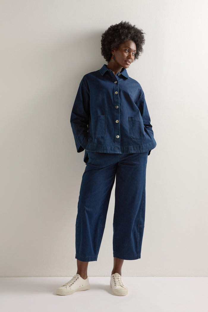 A person in a Stretch Denim Jacket with a dark blue boxy silhouette and matching pants stands against a white wall, looking aside, hands in pockets.