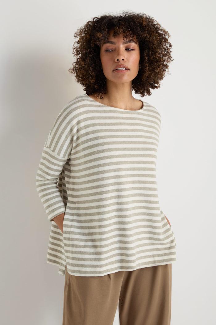 A woman stands against a plain white background wearing the Stripe Flared Fleece Top with long sleeves and flared stripes, paired with tan wide-leg pants.