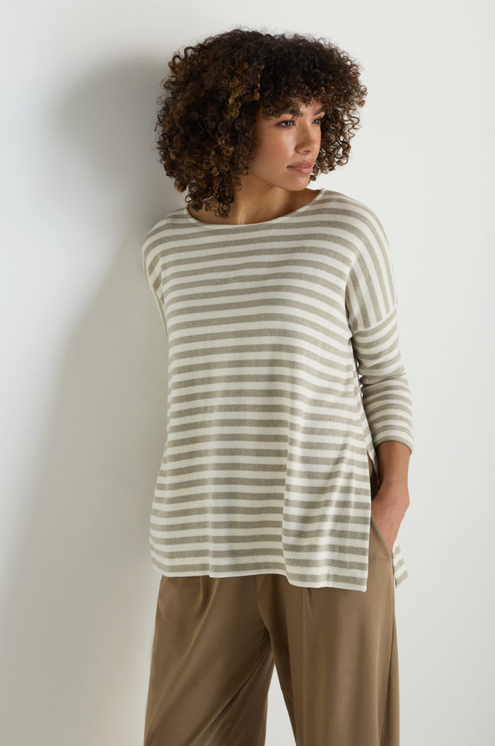 A woman stands against a white wall wearing the Stripe Flared Fleece Top featuring beige and white stripes, paired with tan pants. She has her hands in her pockets and looks to the side.