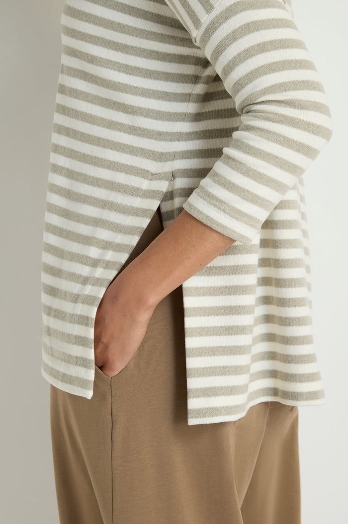 A person wears the Stripe Flared Fleece Top, featuring beige and white stripes, flared silhouette, side slits, and a comfy cotton blend fleece—styled with tan pants and shown from the side with one hand in a pocket.