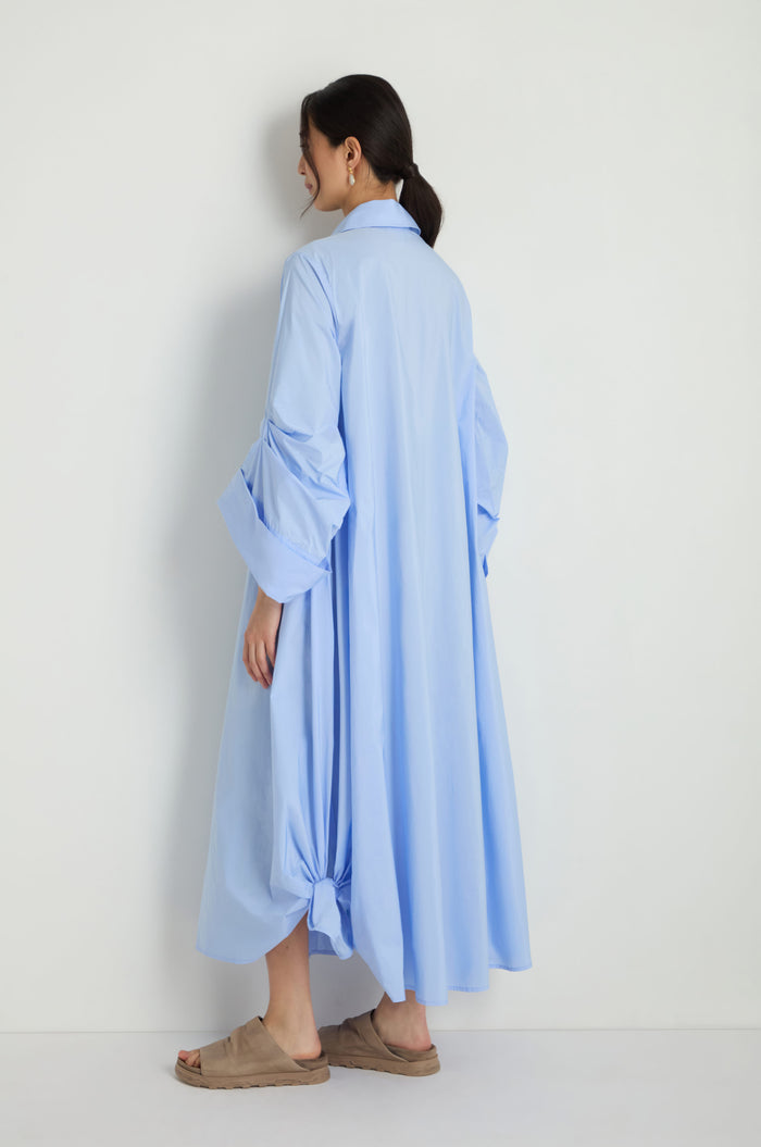 A woman stands against a white wall, partially turned from the camera, wearing the Taffeta Button-Through Sky Dress—a long, light blue dress with rolled sleeves—and tan sandals.