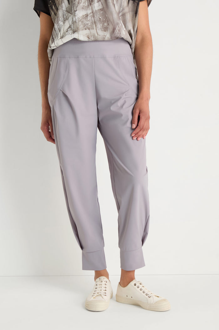 Woman stands against a white wall wearing a white graphic t-shirt, Tapered Cuff Trousers in light gray with pleats, and white sneakers. Her hands are in her pockets as she looks to the side.