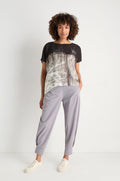 Wearing Tapered Cuff Trousers in light gray stretch fabric, paired with white sneakers and a black and white patterned shirt, the person stands against a plain white background.
