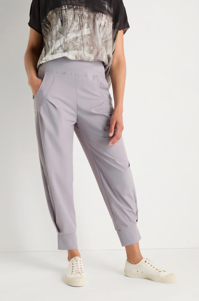 A person wearing light gray Tapered Cuff Trousers with pleated side pockets and a black and white patterned top, standing with hands in pockets.