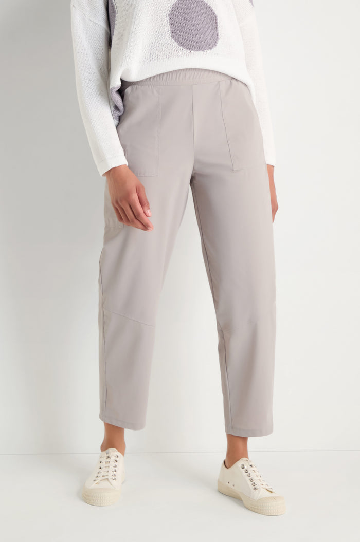 A person models the Tapered Trousers in light gray stretch fabric, paired with a white sweater featuring a large gray dot and off-white sneakers, standing against a plain white background.