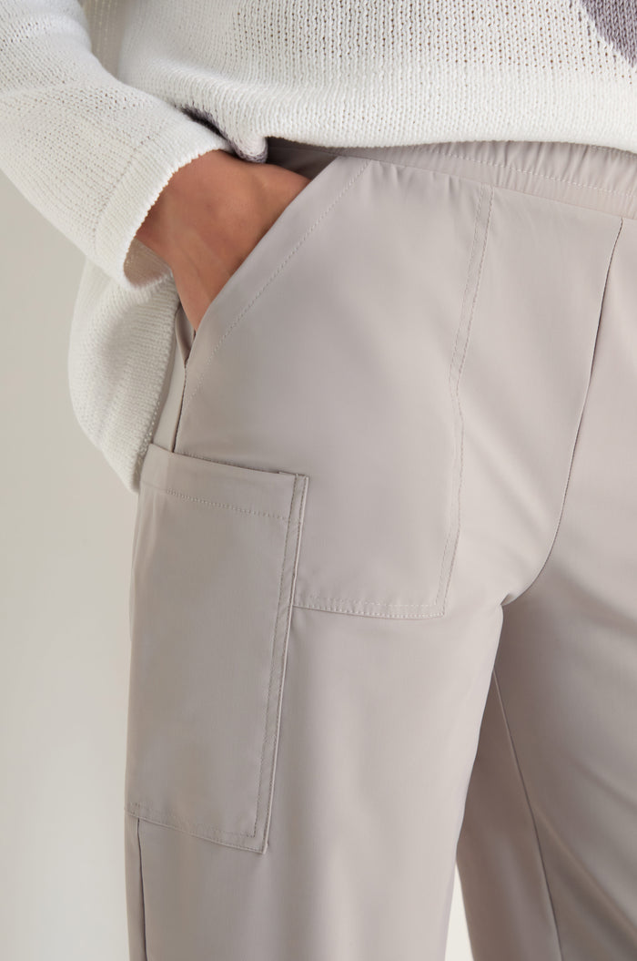 Person wearing Tapered Trousers in light beige with a side pocket and stretch fabric for comfort, styled with a white textured sweater. The trousers have a modern, cropped cut and the person has one hand in the pocket.