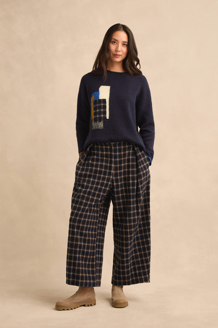 A woman stands against a beige background, wearing Tartan Pleated Trousers with a wide cropped leg in navy plaid, paired with a navy sweater featuring an abstract design and tan boots.