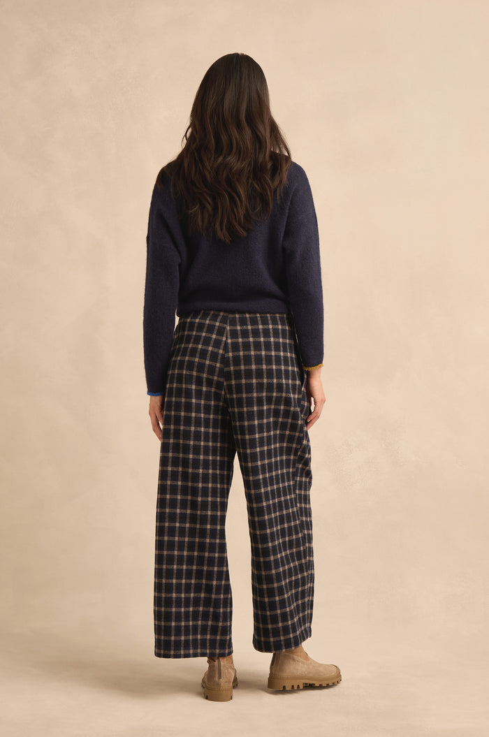A person with long brown hair is shown from behind against a neutral background, wearing a navy sweater, tan boots, and the Tartan Pleated Trousers, which feature a wide cropped leg.