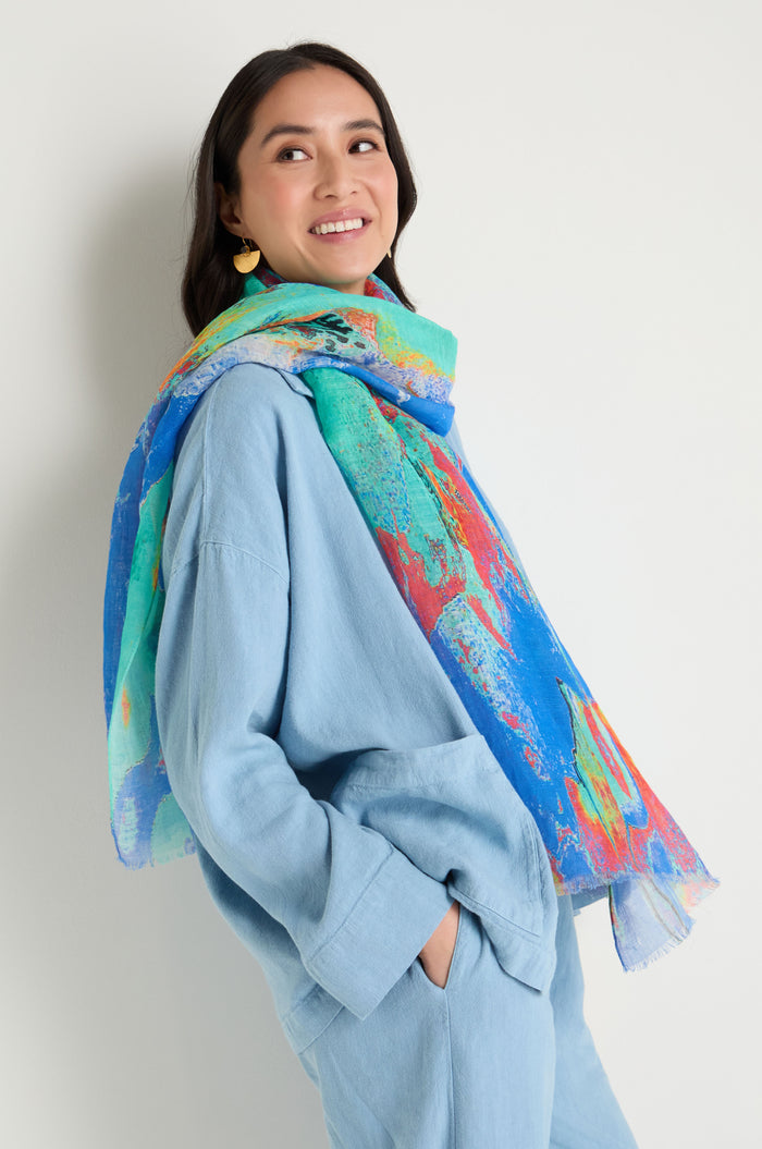 A woman smiles and looks to the side, standing with hands in pockets against a plain background, wearing a light blue outfit paired with the Terra Mare Cotton Linen Scarf.