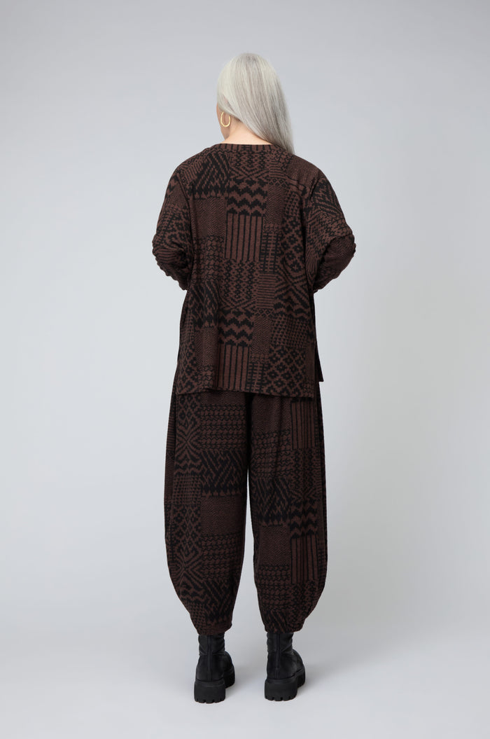 A person with long gray hair stands facing away, wearing an oversized Textured Ikat Patchwork Jersey Top and matching pants with black boots against a plain light background.