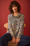 A woman with curly hair sits on a wooden chair, wearing the Textured Prism Jersey Top—a multicolored patterned jersey with a boxy silhouette—paired with navy pants, in front of a red background.
