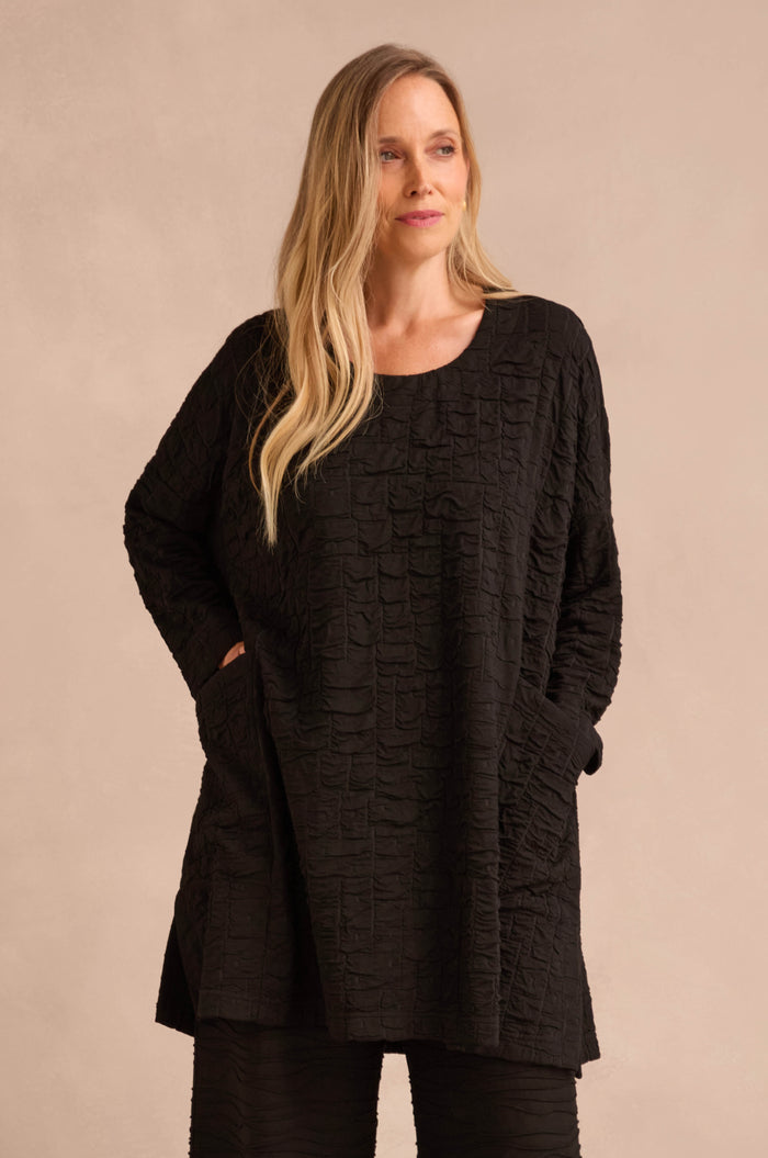 A woman wearing the Textured Squares Jersey Tunic, which features long sleeves, pockets, and a subtle square pattern, stands against a neutral background.