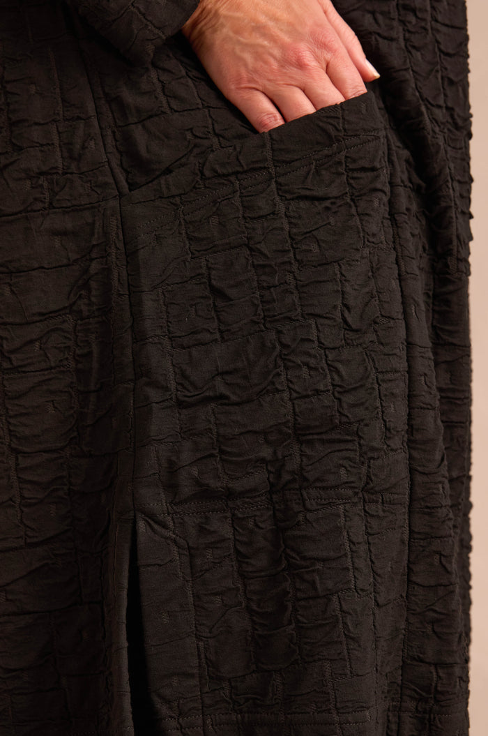 A hand is partially placed inside the pocket of a Textured Squares Jersey Tunic, highlighting the garment’s crinkled pattern in close-up detail.