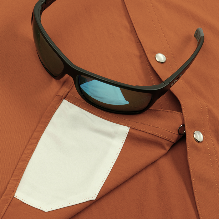 Close-up of a rust-colored shirt with white pocket and black sunglasses resting on it, showcasing The 2005 shirt details.