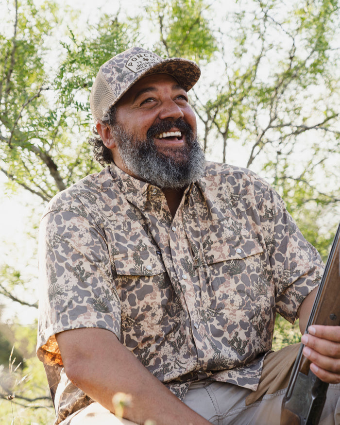 Bearded man outdoors wearing a patterned short-sleeve shirt called The Abilene, smiling and holding a rifle.