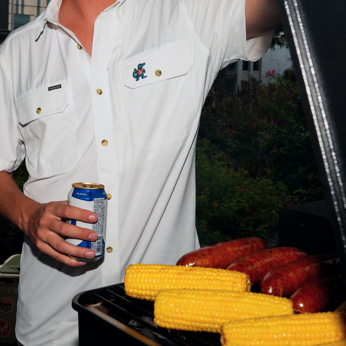 Person wearing The Albert white shirt grilling corn and sausages while holding a drink outdoors