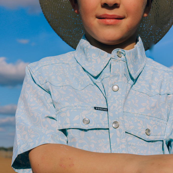 Close-up of a child wearing The Alma light blue outdoor shirt with floral print and silver snaps against a blue sky background