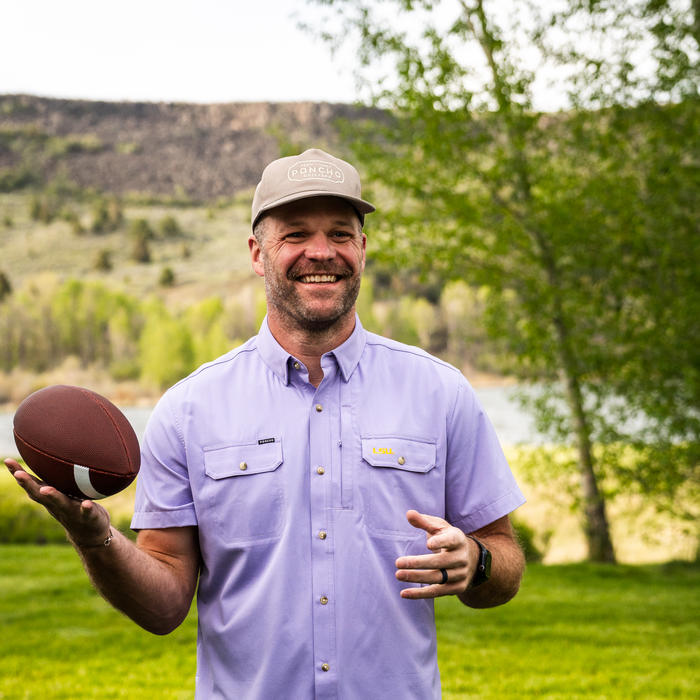 Man smiling outdoors wearing The Baton Rouge shirt while holding a football in a natural green landscape