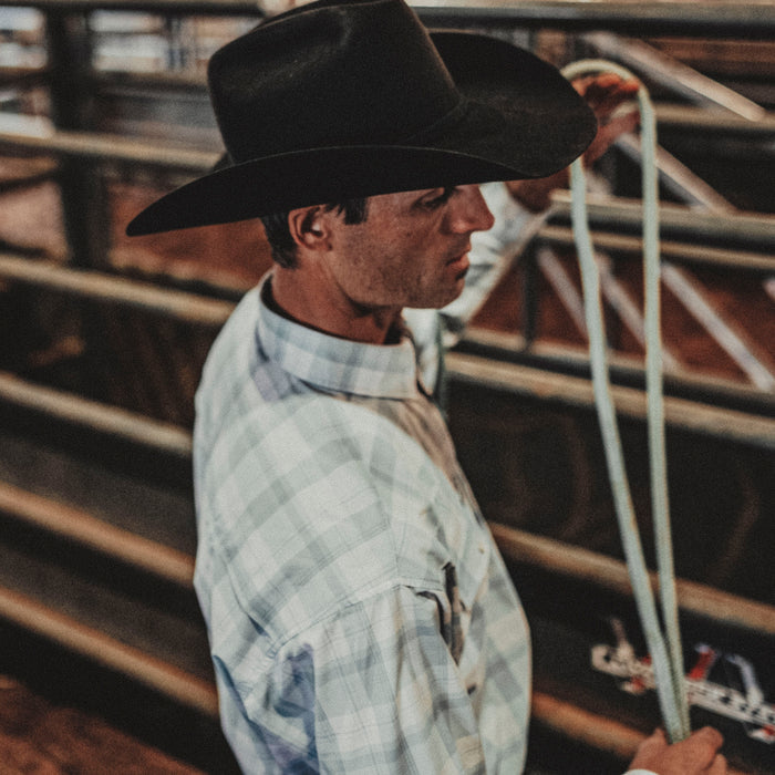 Man wearing The Billings outdoor shirt and black wide-brim hat handling rope in rustic setting