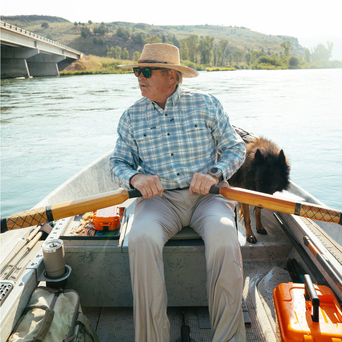 Man wearing The Blue Bison shirt rowing a boat on a river with a dog and outdoor gear nearby