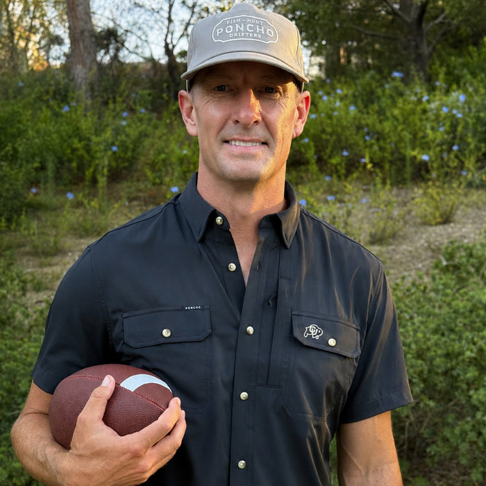 Man wearing The Buff shirt holding a football outdoors with trees and grass in the background