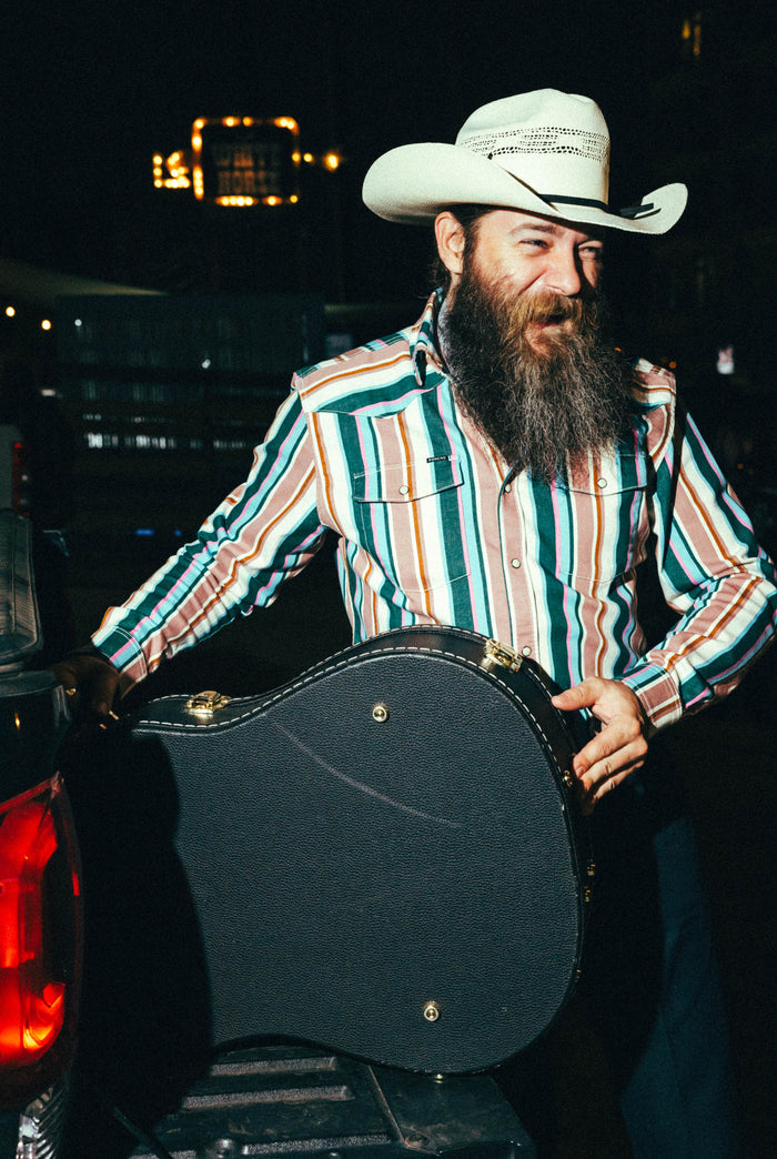 Man wearing The Frontman striped outdoor shirt and cowboy hat holding guitar case at night