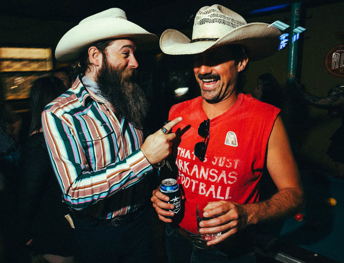 Two men wearing cowboy hats, one in a colorful striped shirt and the other in a sleeveless red shirt labeled The Frontman, laughing and holding drinks indoors