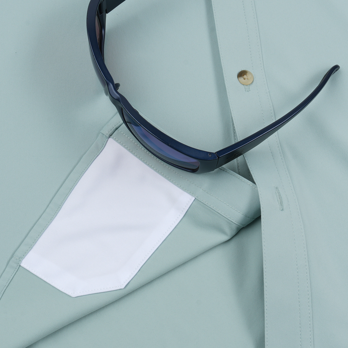 Close-up of a light green shirt with a white pocket and dark blue sunglasses resting on it.