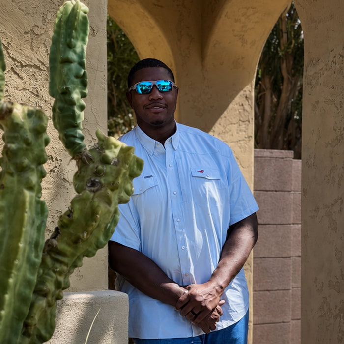 Man wearing The Pony Up shirt and blue reflective sunglasses standing next to a cactus outdoors