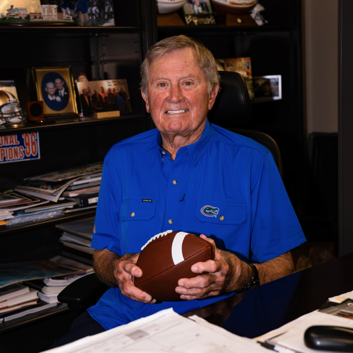 Older man smiling while sitting at desk holding a football wearing The Swamp Chomp blue shirt in an office setting