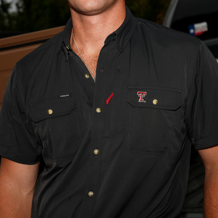 Close-up of a man wearing a black short-sleeve button-up shirt featuring The Tortilla logo on the chest pocket