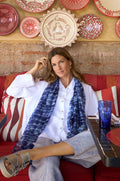 A woman in a white shirt and a Tie Dye Print Scarf sits on a red cushioned bench with patterned pillows, in front of a wall decorated with red and white plates.