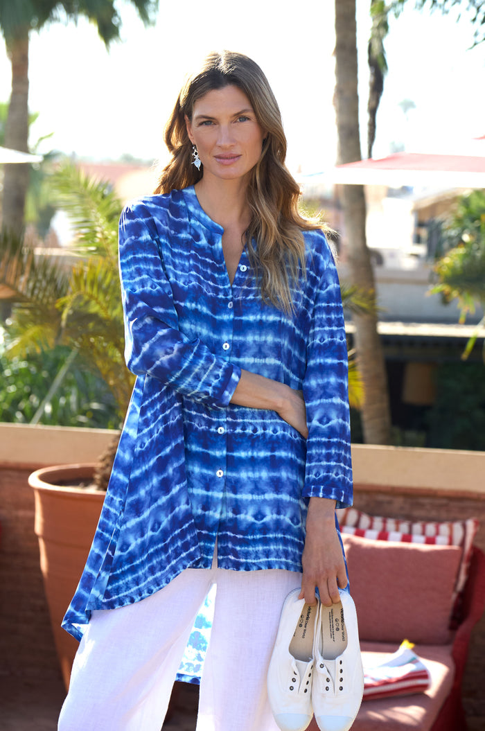 A woman stands outdoors among palm trees and patio furniture, wearing the Tie Dye Printed Viscose Shirt in blue and white with white pants, holding white shoes in one hand.