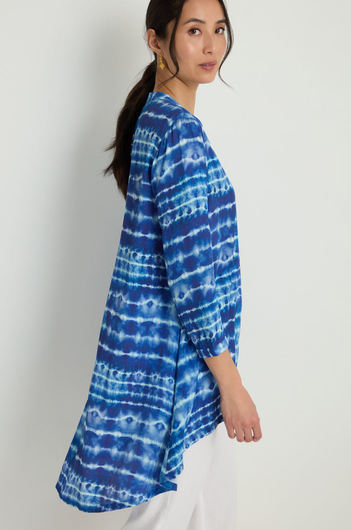A woman stands sideways against a plain light background, wearing the Tie Dye Printed Viscose Shirt with long sleeves in blue and white, paired with white pants.