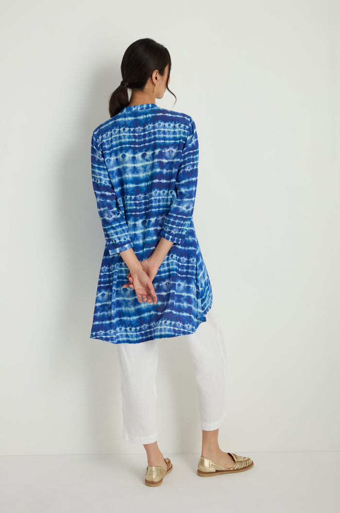 A woman faces a white wall, wearing the Tie Dye Printed Viscose Shirt styled as a tunic with white pants and gold shoes, her hands clasped behind her back.