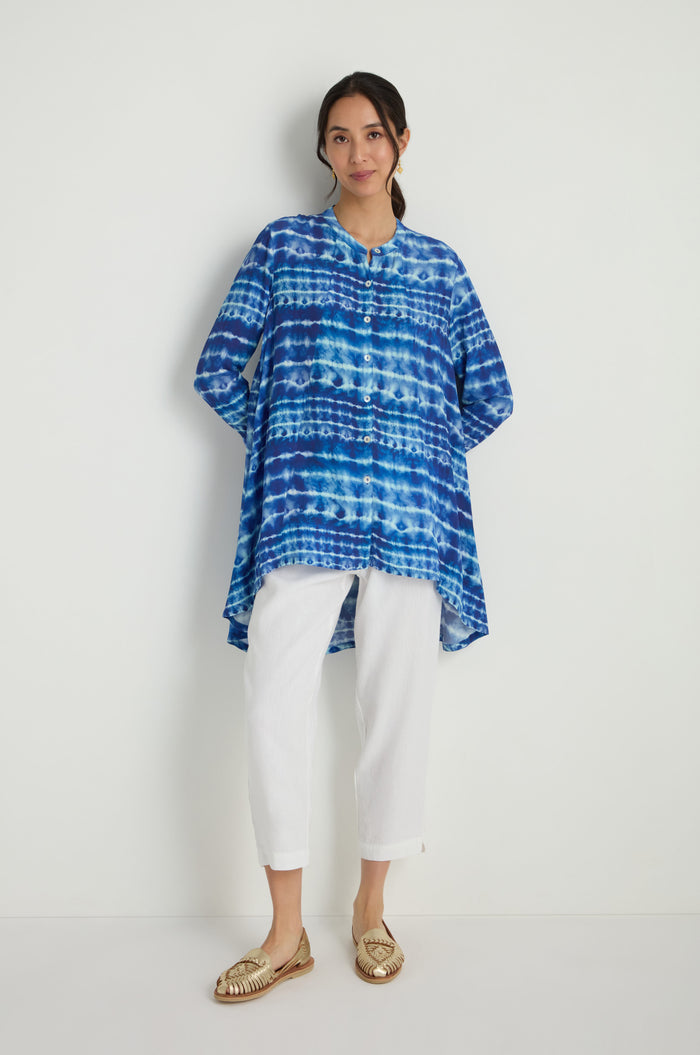 A woman stands against a white wall wearing a Tie Dye Printed Viscose Shirt, white pants, and gold woven shoes.