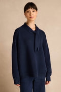 A woman stands against a beige background, wearing the Tie Neck Navy Ribbed Knit sweater paired with dark pants. She looks directly at the camera with a neutral expression, while the relaxed fit and collar detail of the jumper add effortless sophistication to her look.