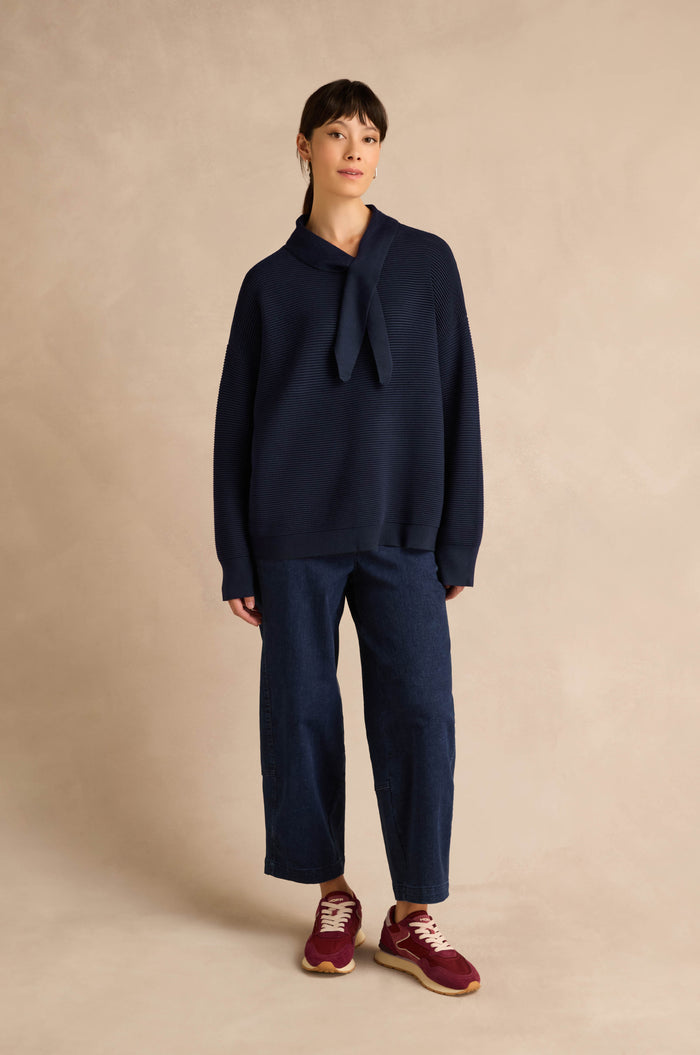 A woman stands against a beige textured background wearing the Tie Neck Navy Ribbed Knit jumper with a relaxed fit, paired with wide-leg dark blue pants and red sneakers.
