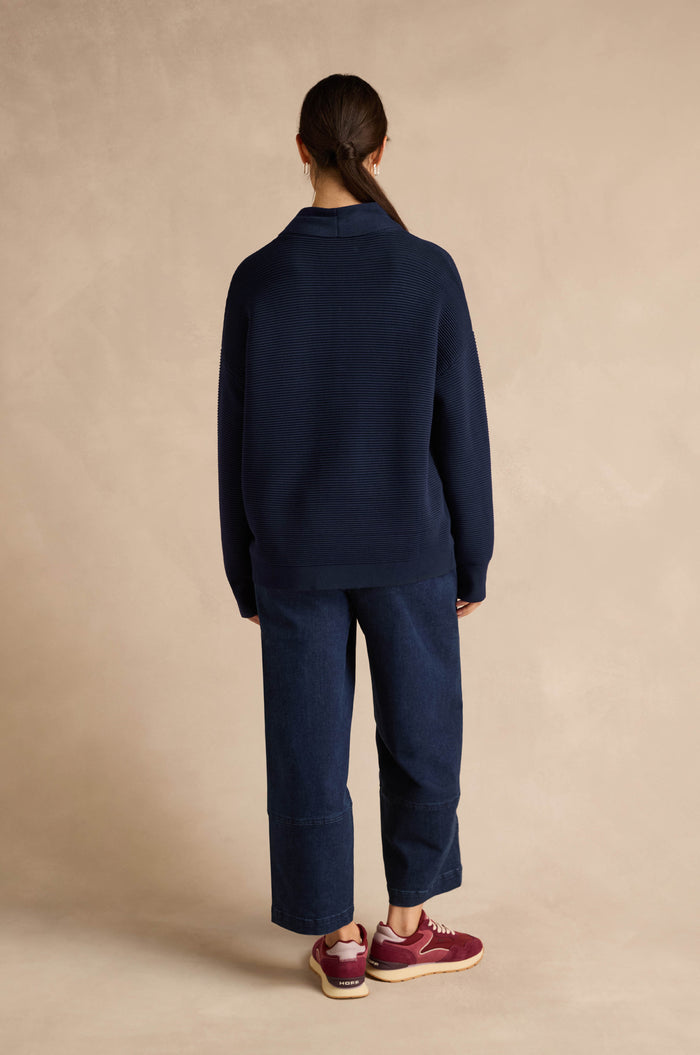 A person stands with their back to the camera against a beige background, wearing the Tie Neck Navy Ribbed Knit in a relaxed fit, paired with wide-leg jeans and maroon sneakers. Their hair is tied back in a low ponytail.